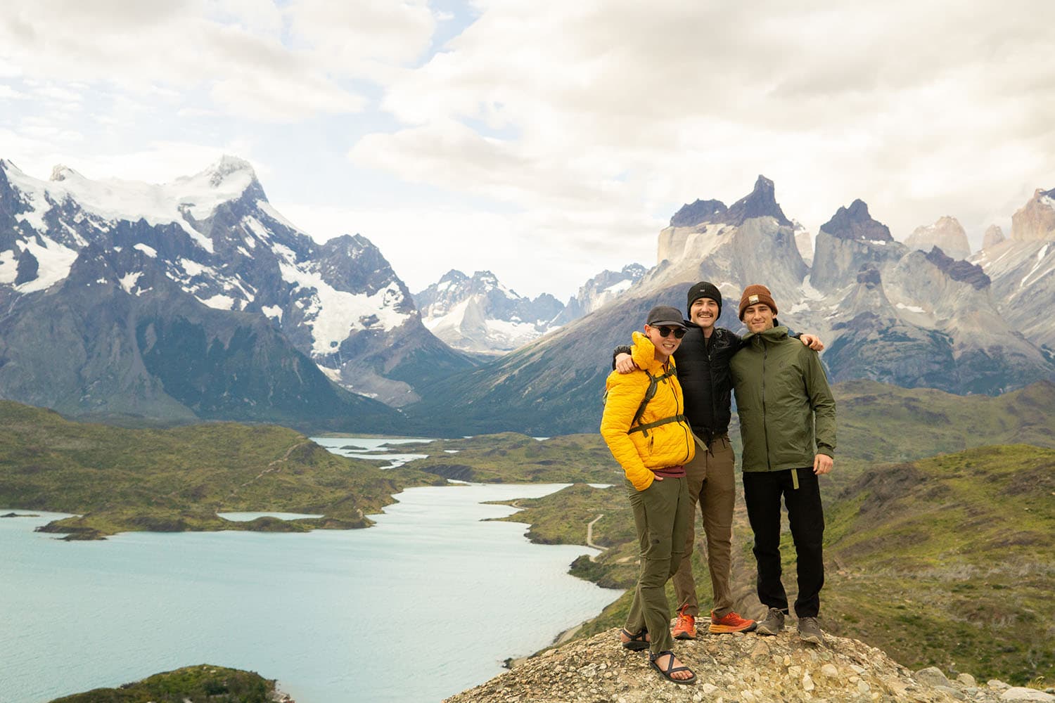 Three friends arm in arm on a rock above a turquoise lake, with snow-capped peaks behind them