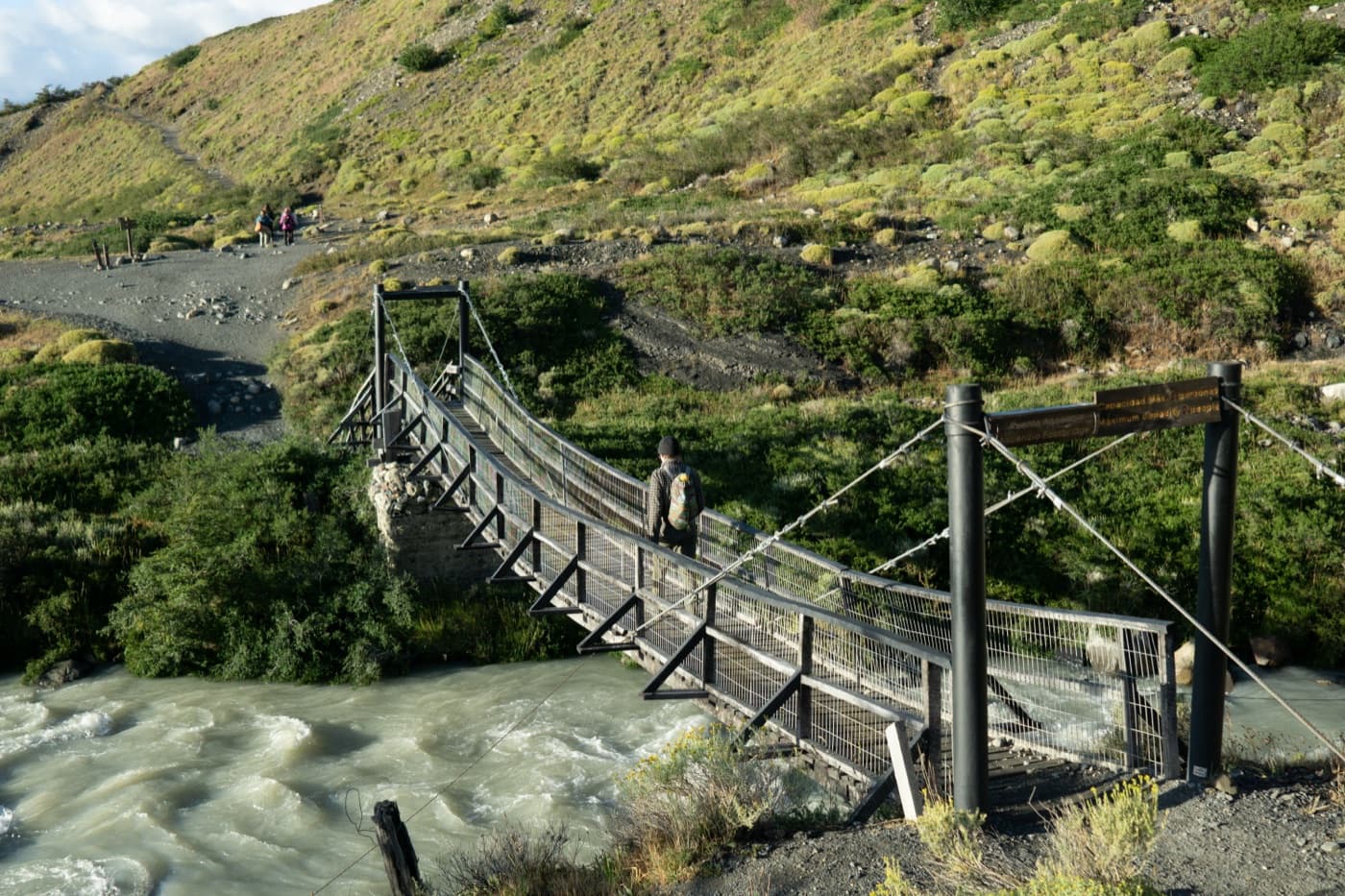 A hiker crossing a suspension footbridge over a rushing glacial river